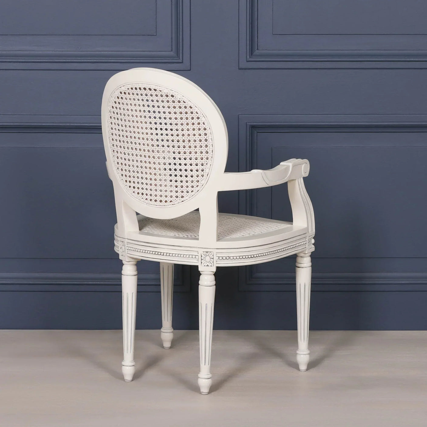 White cane back dining chair with armrests and carved wood details against blue wall