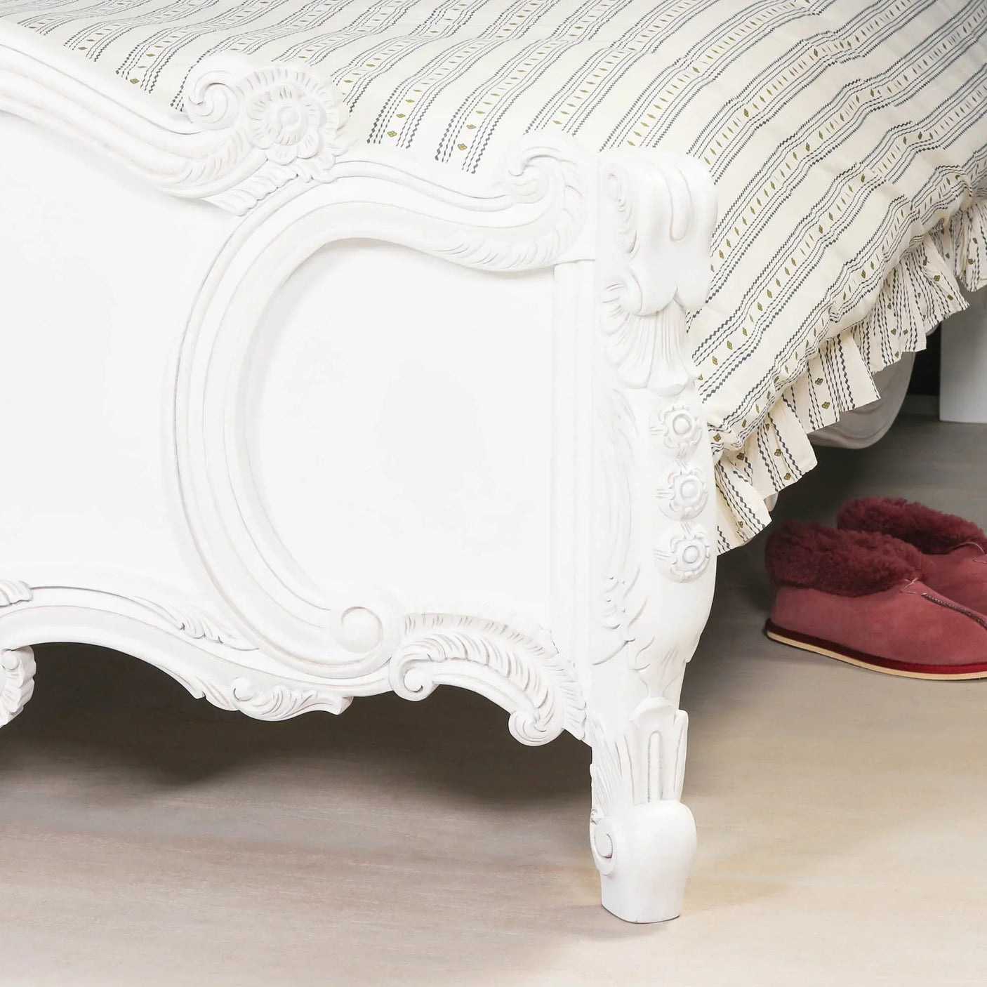 White carved wood bed frame foot with striped bedding and pink slippers on wooden floor