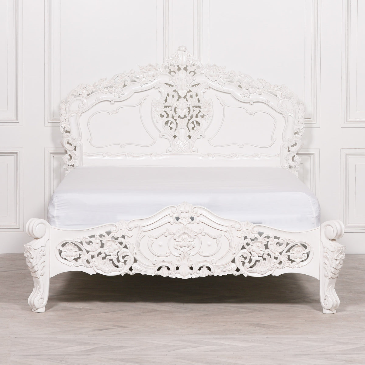 An ornate white carved wooden bed frame with a matching white mattress, set against a white paneled wall and light wood floor.
