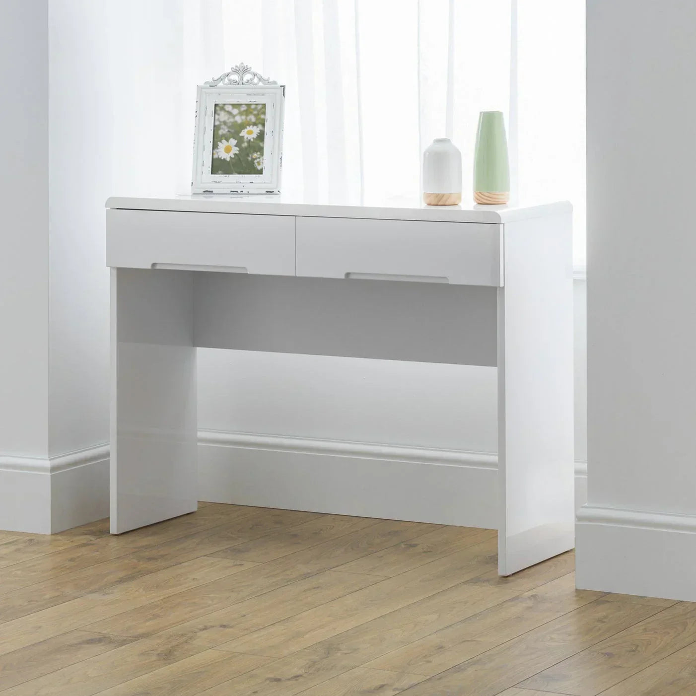 White dressing table with two drawers, vases, and photo frame on wooden floor by window