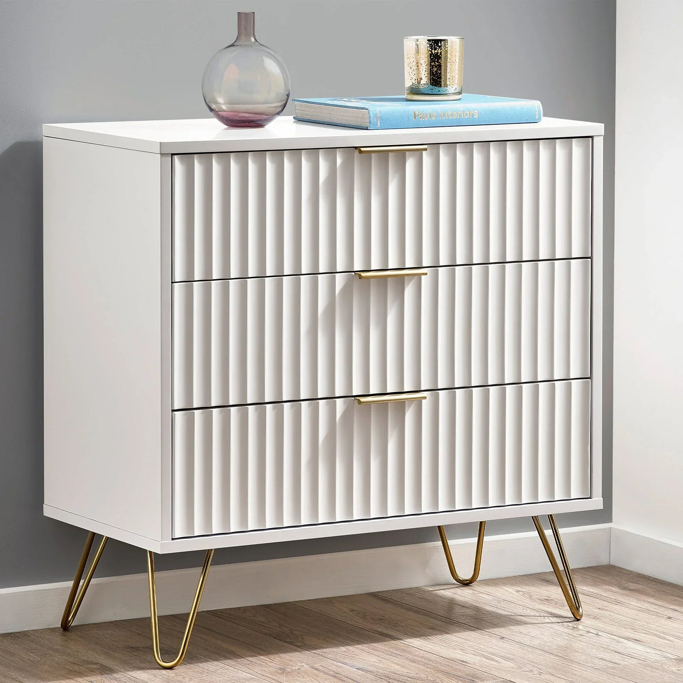 White three drawer chest with ribbed fronts and gold handles on gold hairpin legs