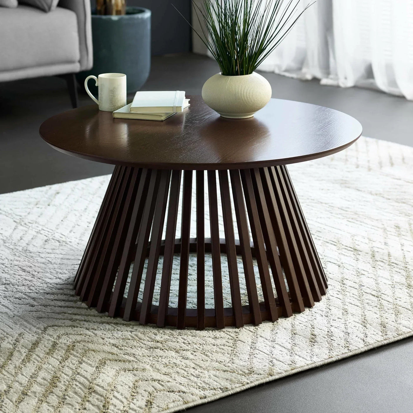 Dark wood round coffee table with slatted base on textured rug, books and vase on top
