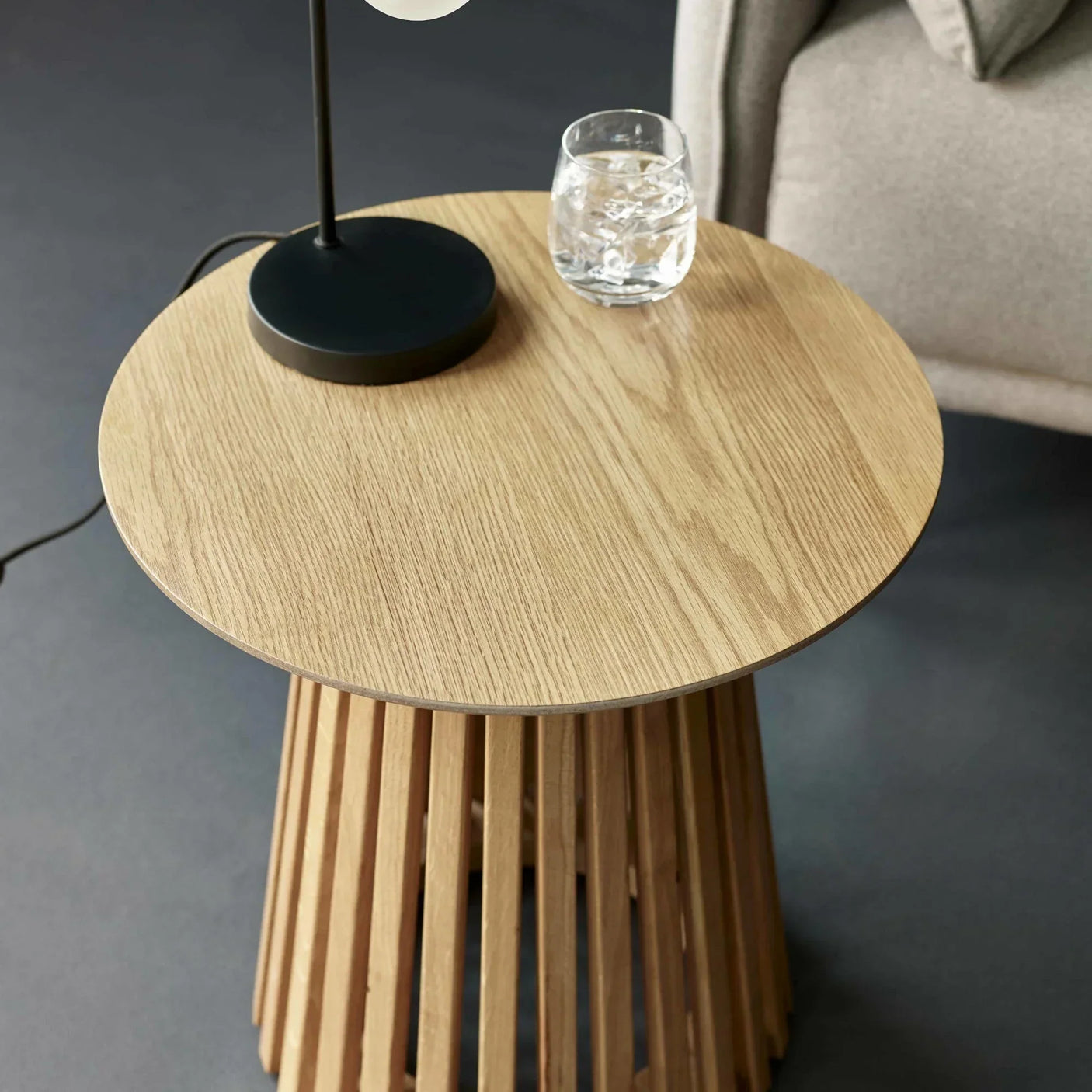 Round oak side table with slatted base beside beige sofa, lamp and glass on top