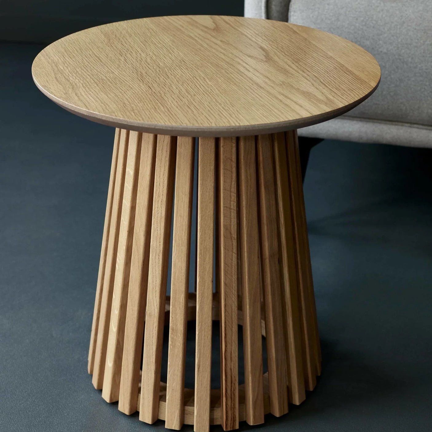 Round oak side table with slatted wooden base beside grey sofa