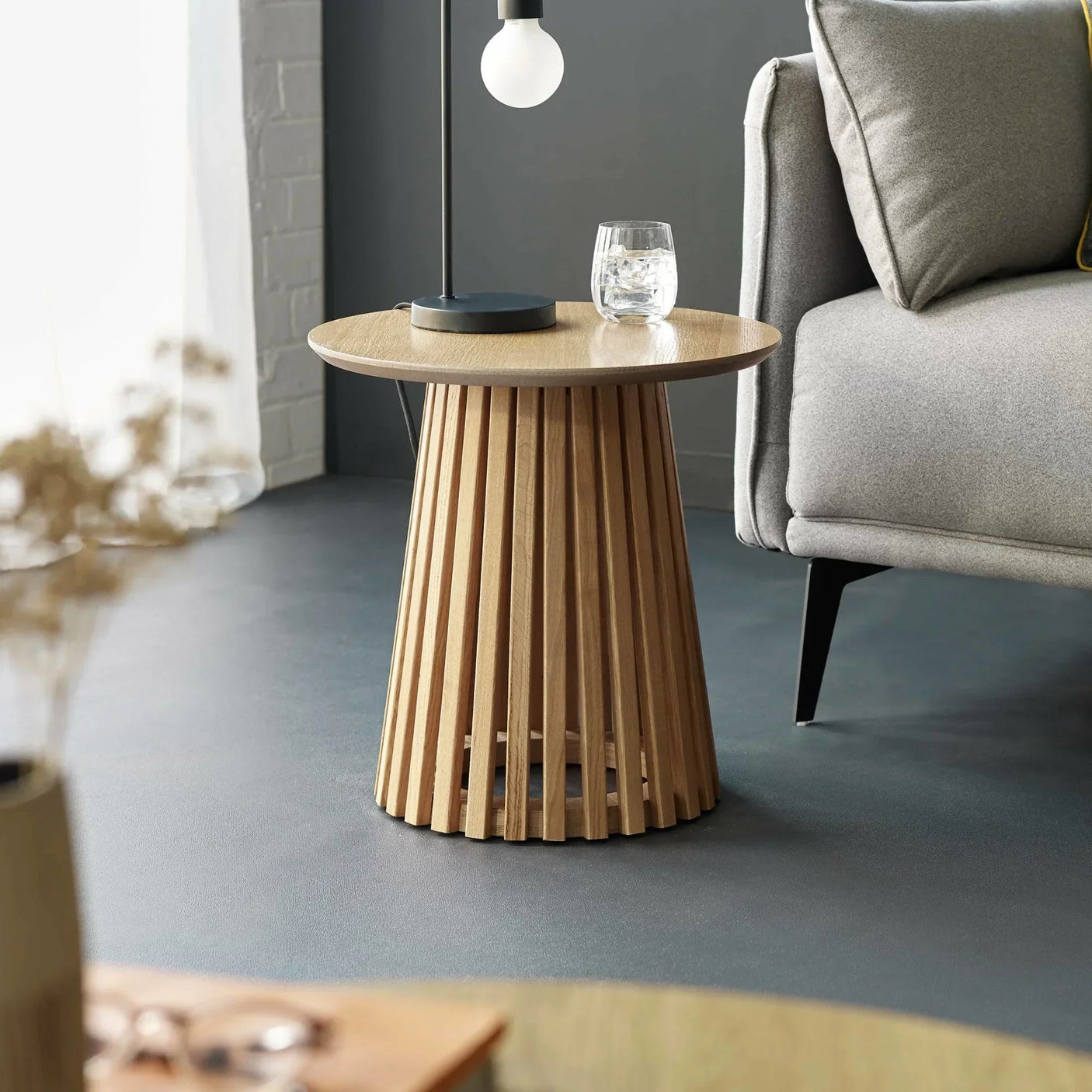 Round oak side table with slatted base beside grey sofa in modern living room