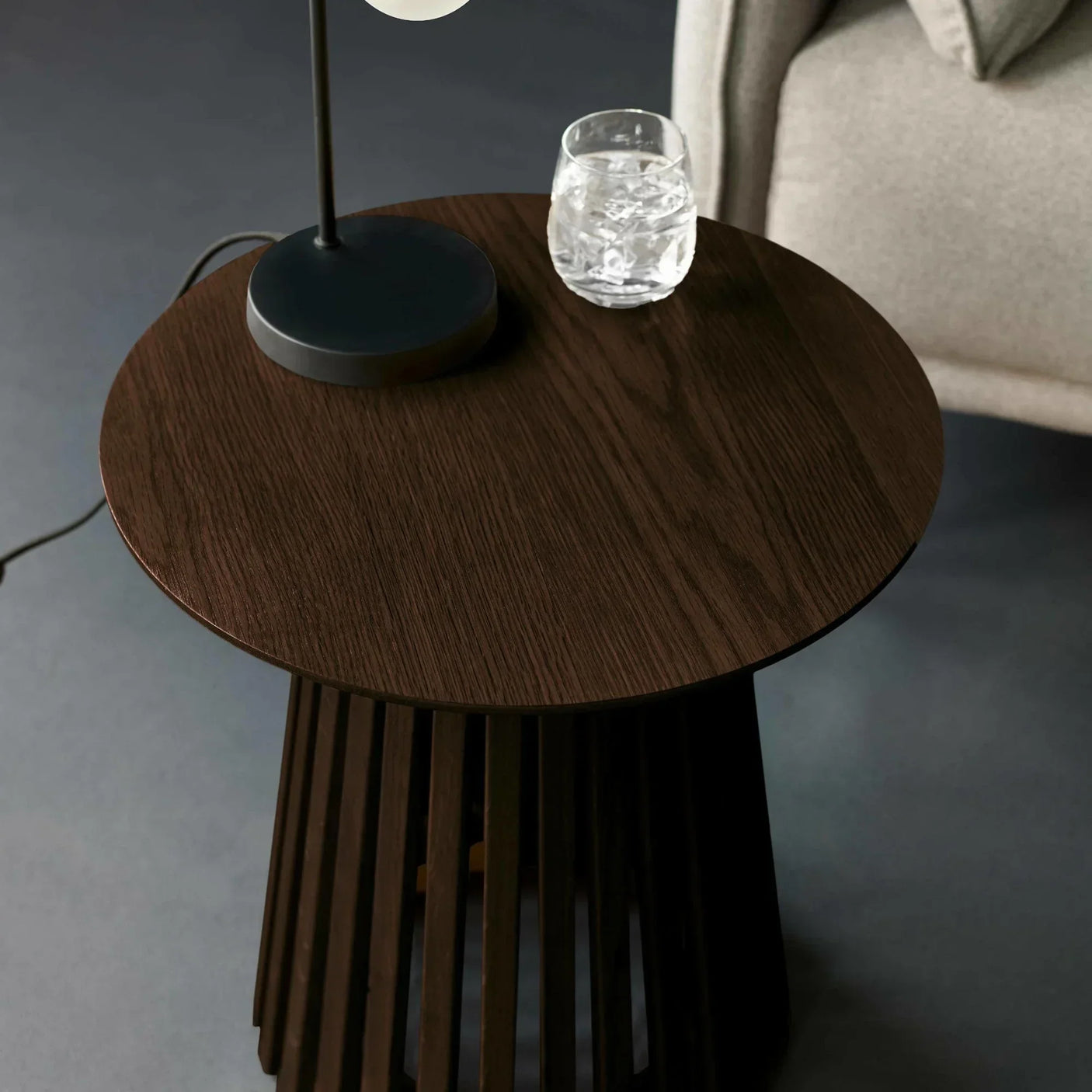 Dark wood side table with modern slatted base, lamp, and glass of water beside sofa