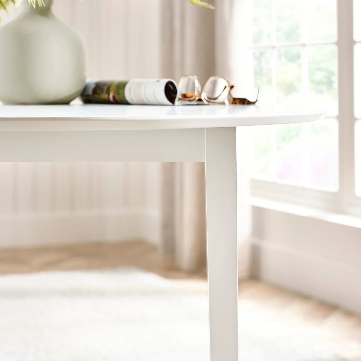 White wooden dining table with tapered leg beside window, vase and magazine on top