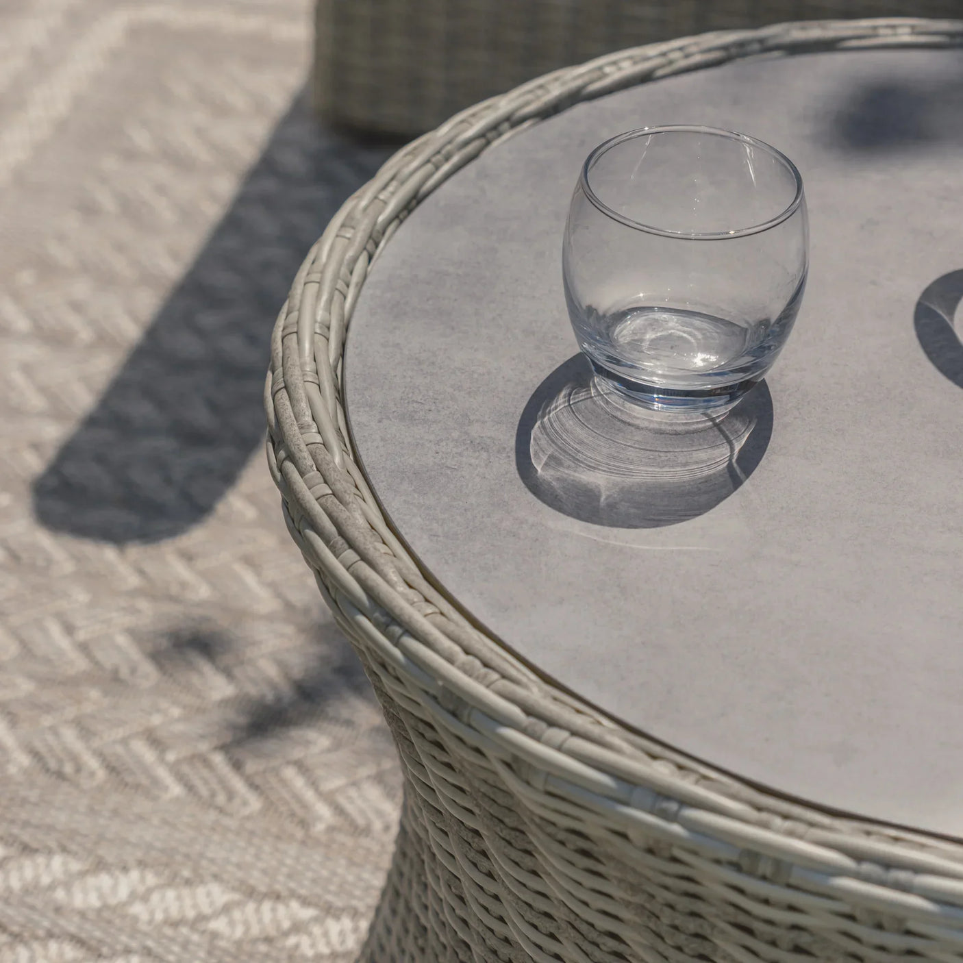 Round rattan outdoor coffee table with glass top and clear tumbler on patterned rug