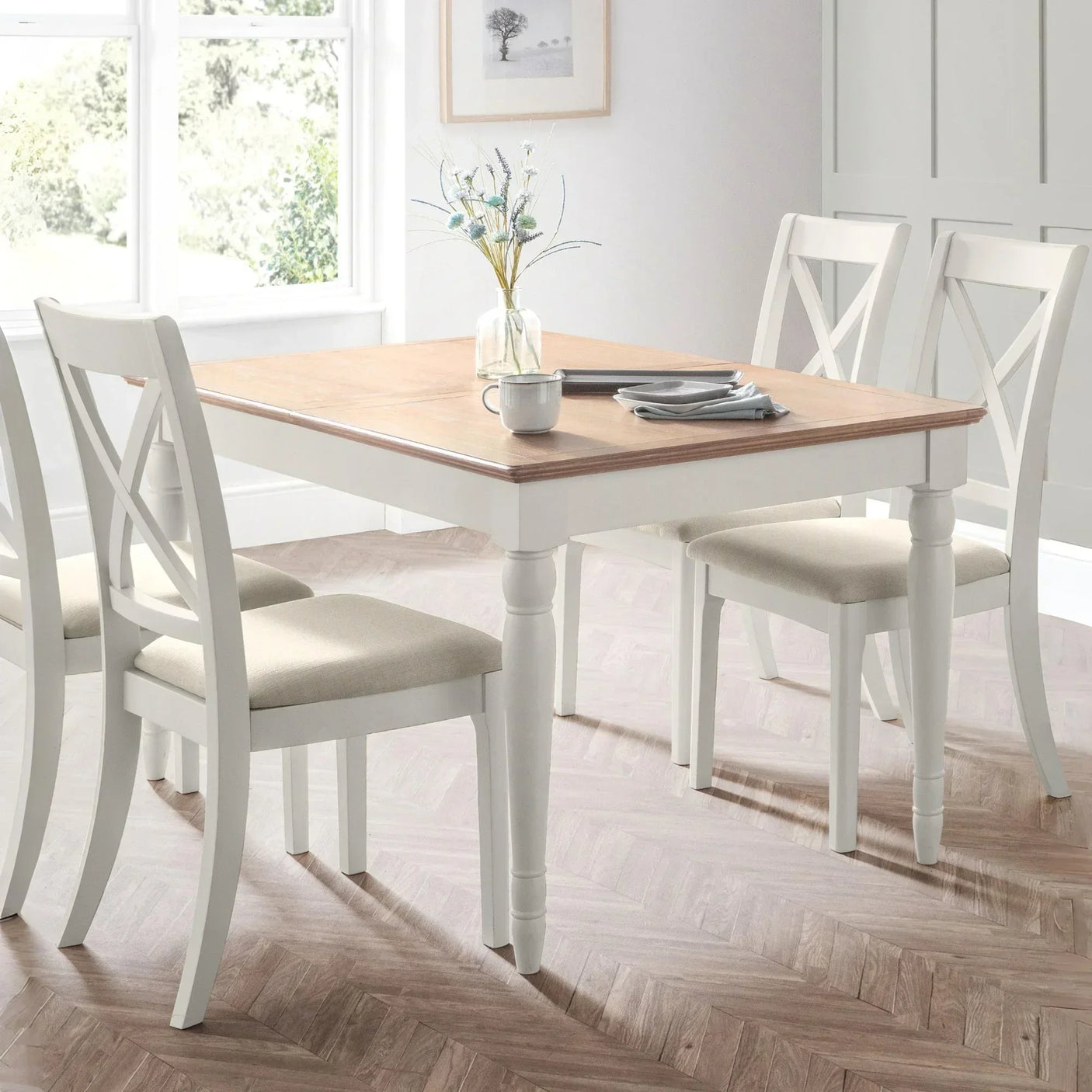 Wooden dining table with white legs and four cushioned chairs in bright dining room