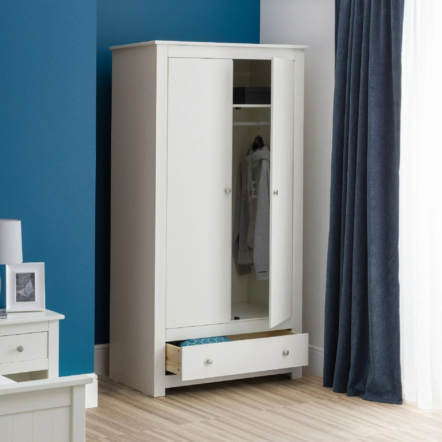 White wooden wardrobe with double doors, hanging rail, and open bottom drawer in bedroom