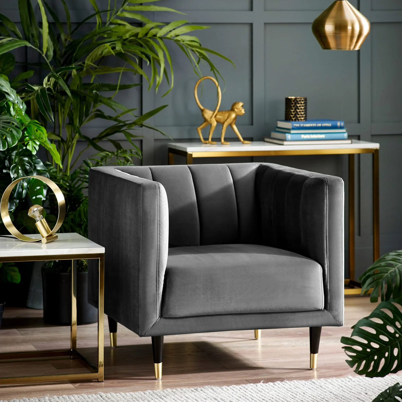 Grey velvet armchair with gold-tipped legs beside plants and modern side tables in living room