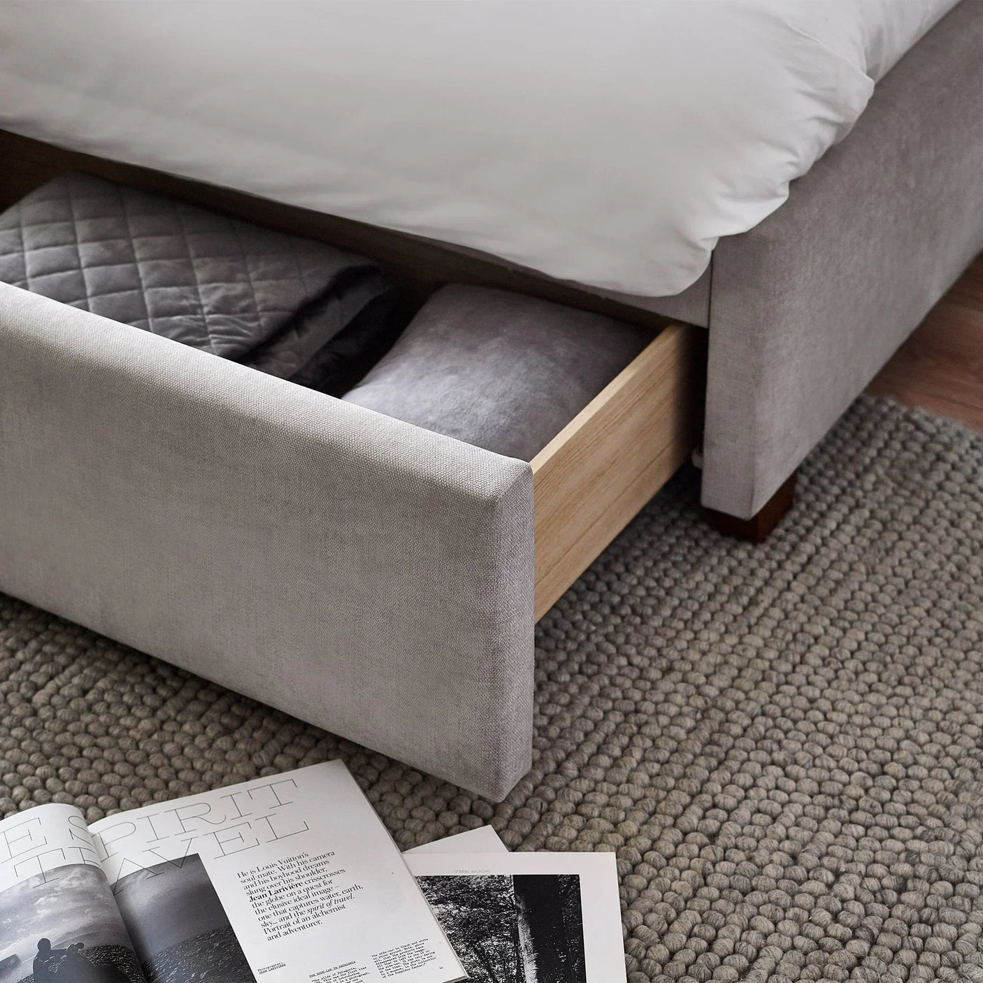 Grey upholstered storage bed with open drawer containing blankets on textured rug