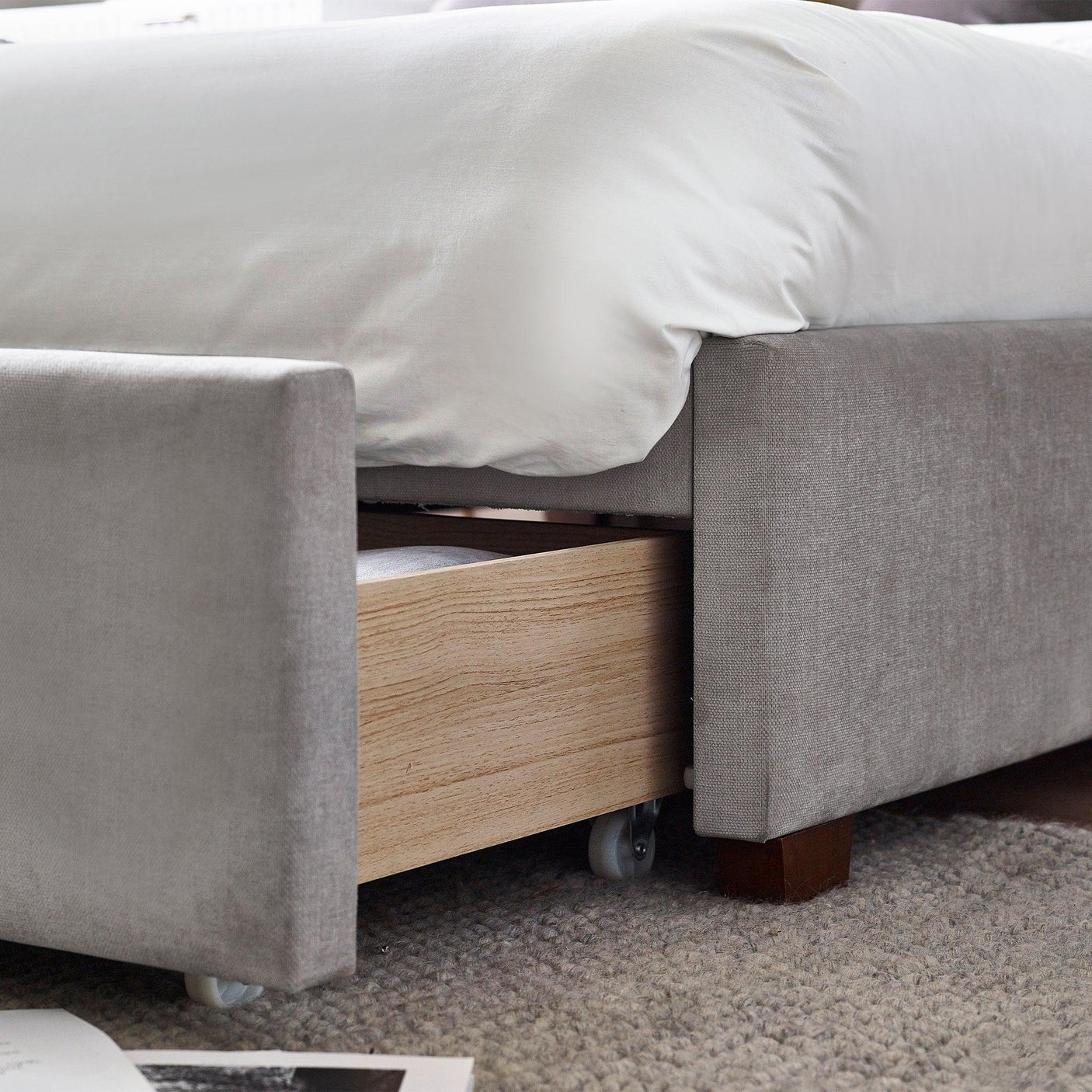 Grey upholstered bed with wooden storage drawer on castors and white bedding