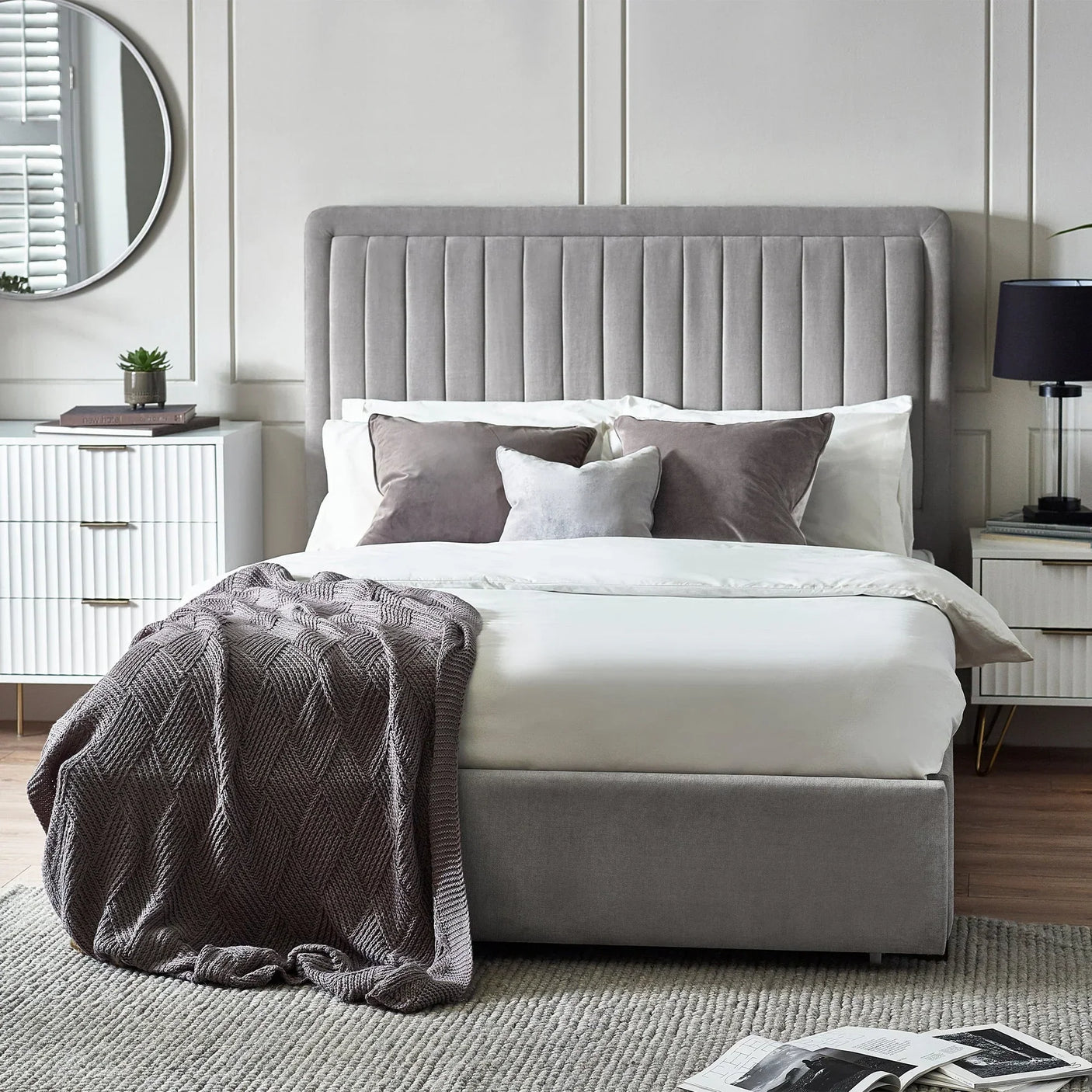 Grey upholstered bed with tall headboard, neatly made with pillows, throw, and bedside tables