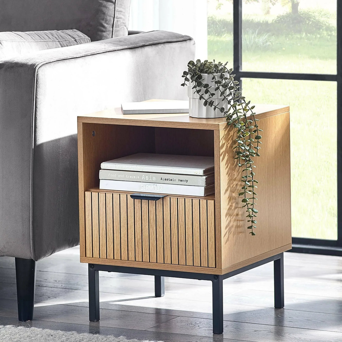 Oak side table with storage shelf, black metal legs, books and potted plant beside sofa