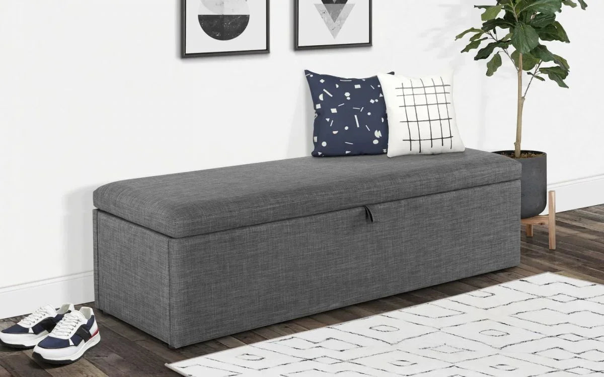 Grey fabric storage bench with lift-up lid, two patterned cushions, and indoor plant
