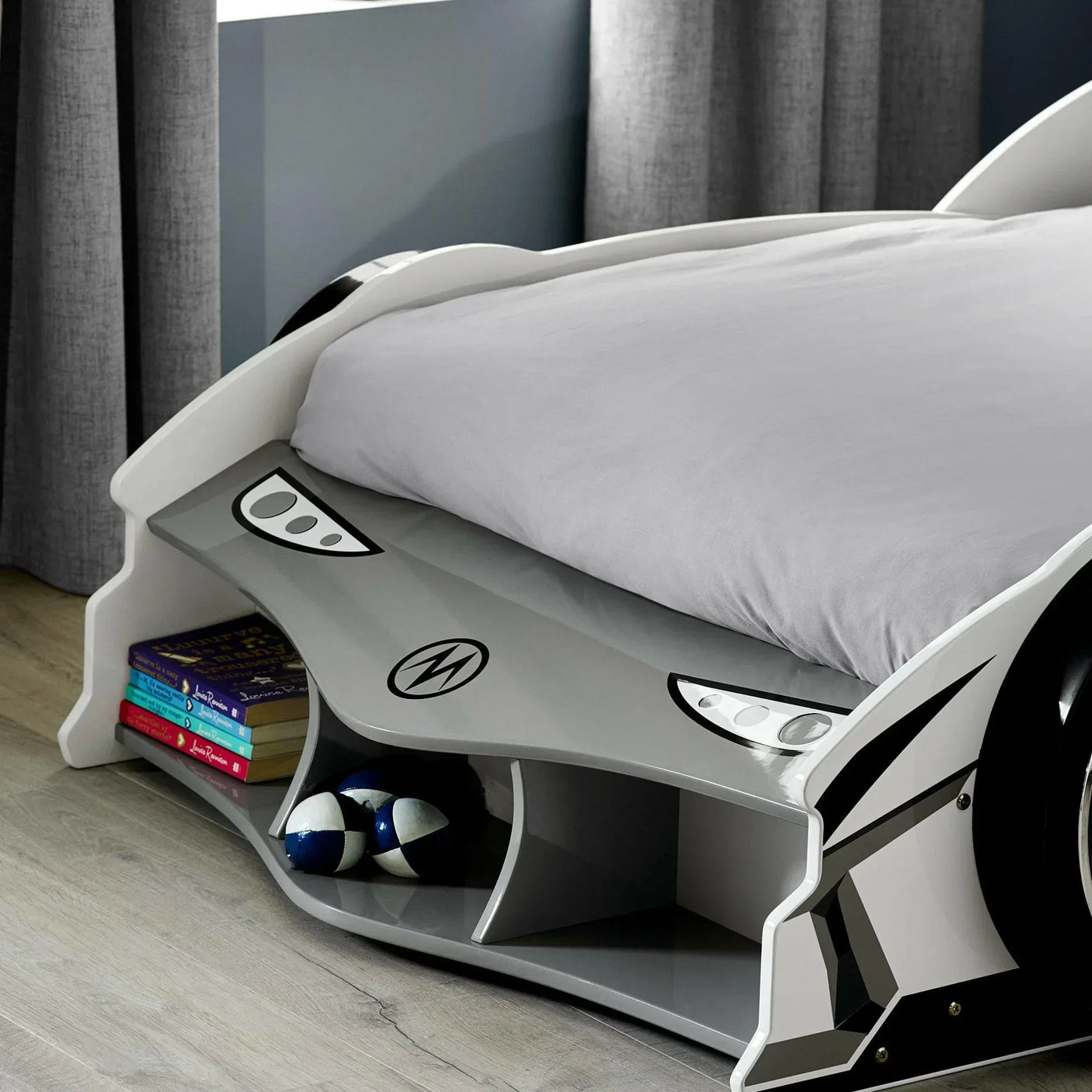 Grey car-shaped kids bed with open storage shelf featuring books and toy balls