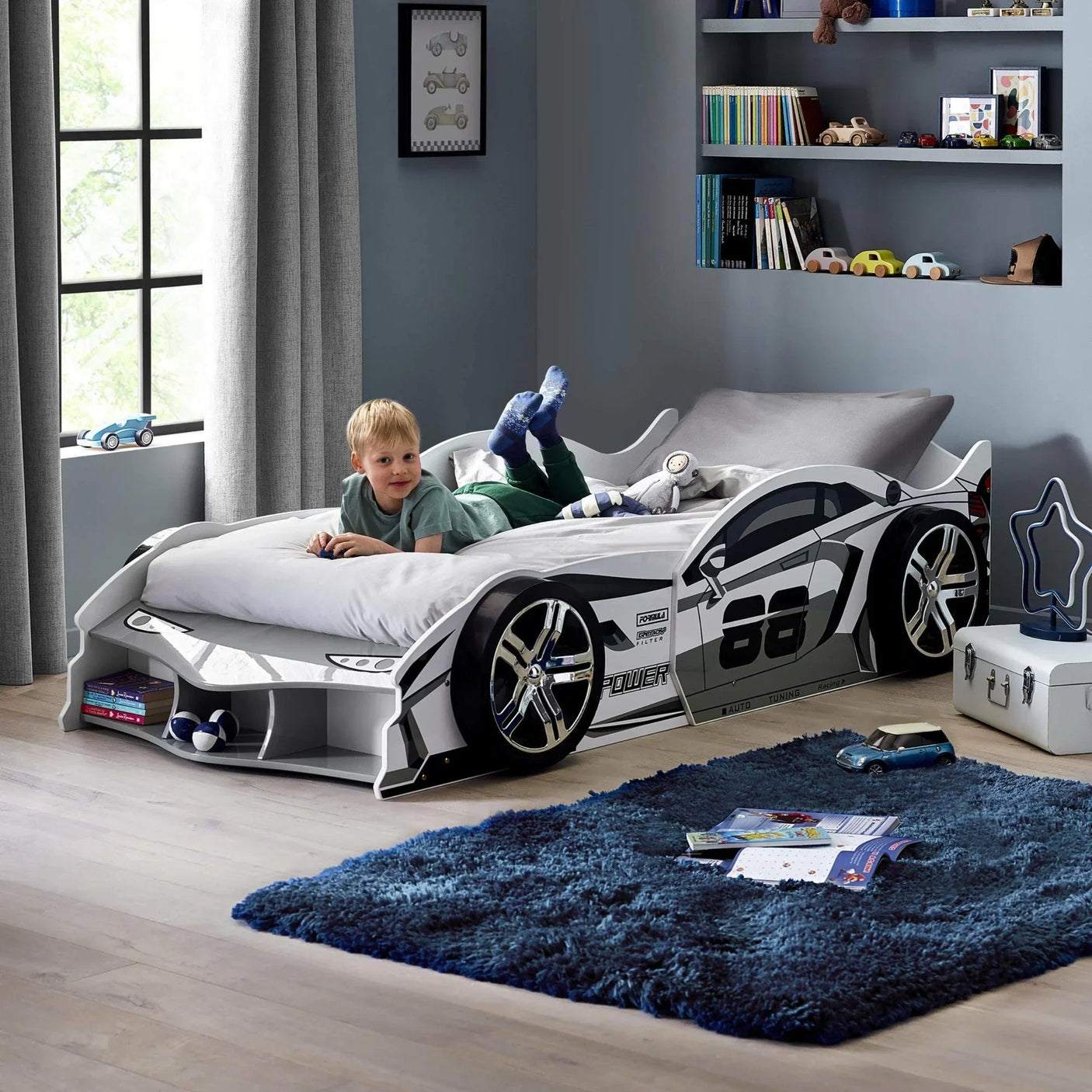 Child lying on white race car bed with storage, in modern kids bedroom with grey walls