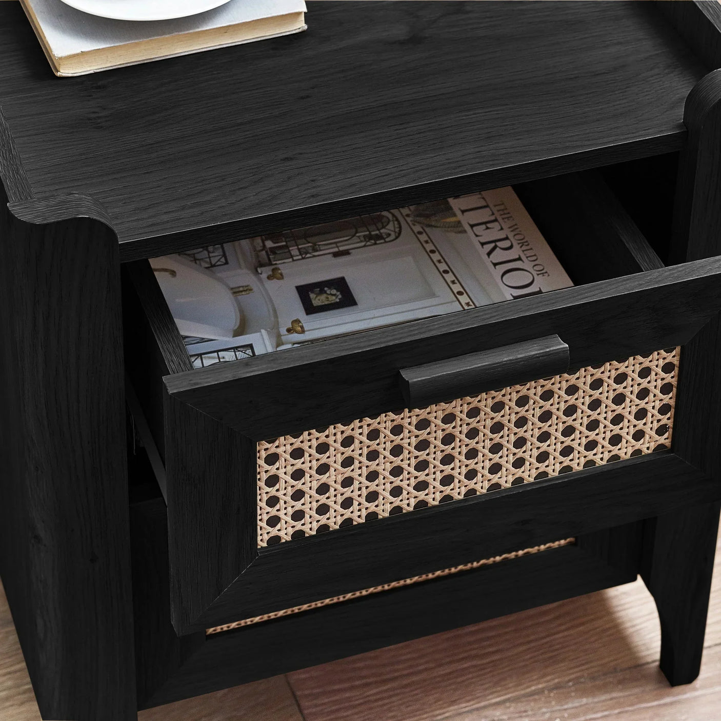 Black rattan effect bedside table with open drawer and magazine inside, wood flooring