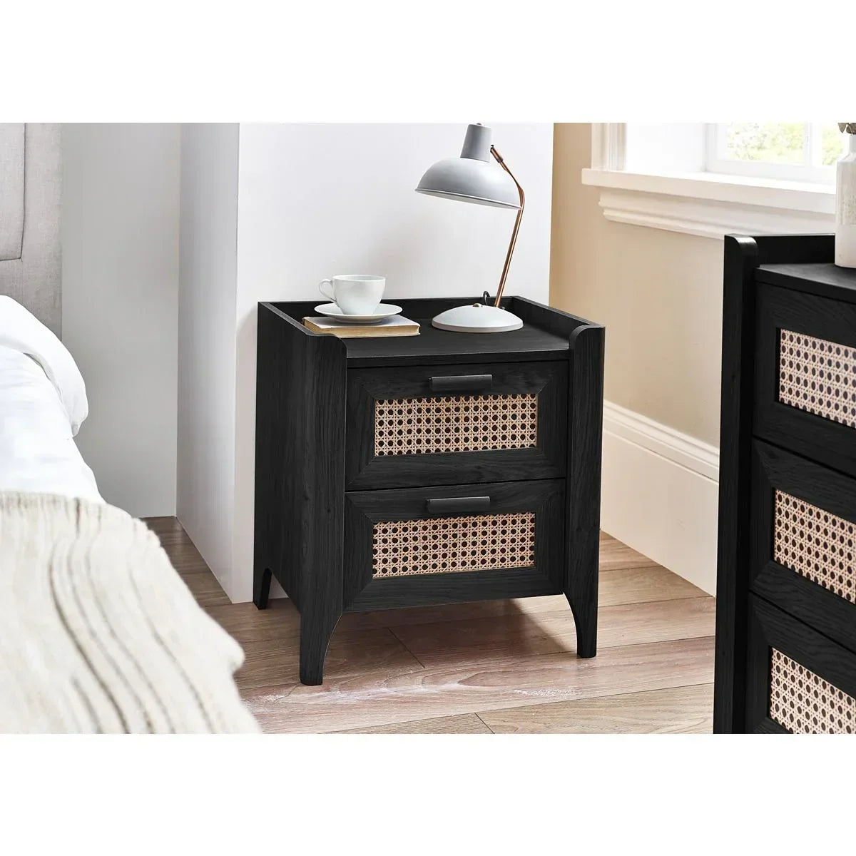 Black wooden bedside table with two rattan drawers, lamp and cup on top, bedroom setting