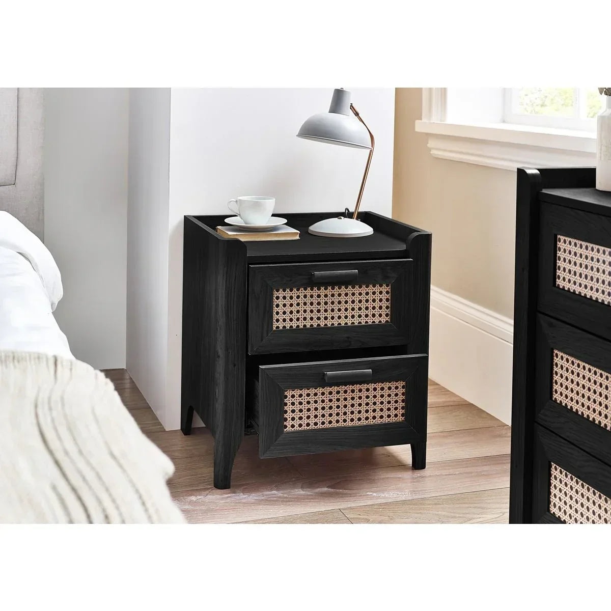 Black wooden bedside table with rattan drawers beside bed, topped with lamp and cup
