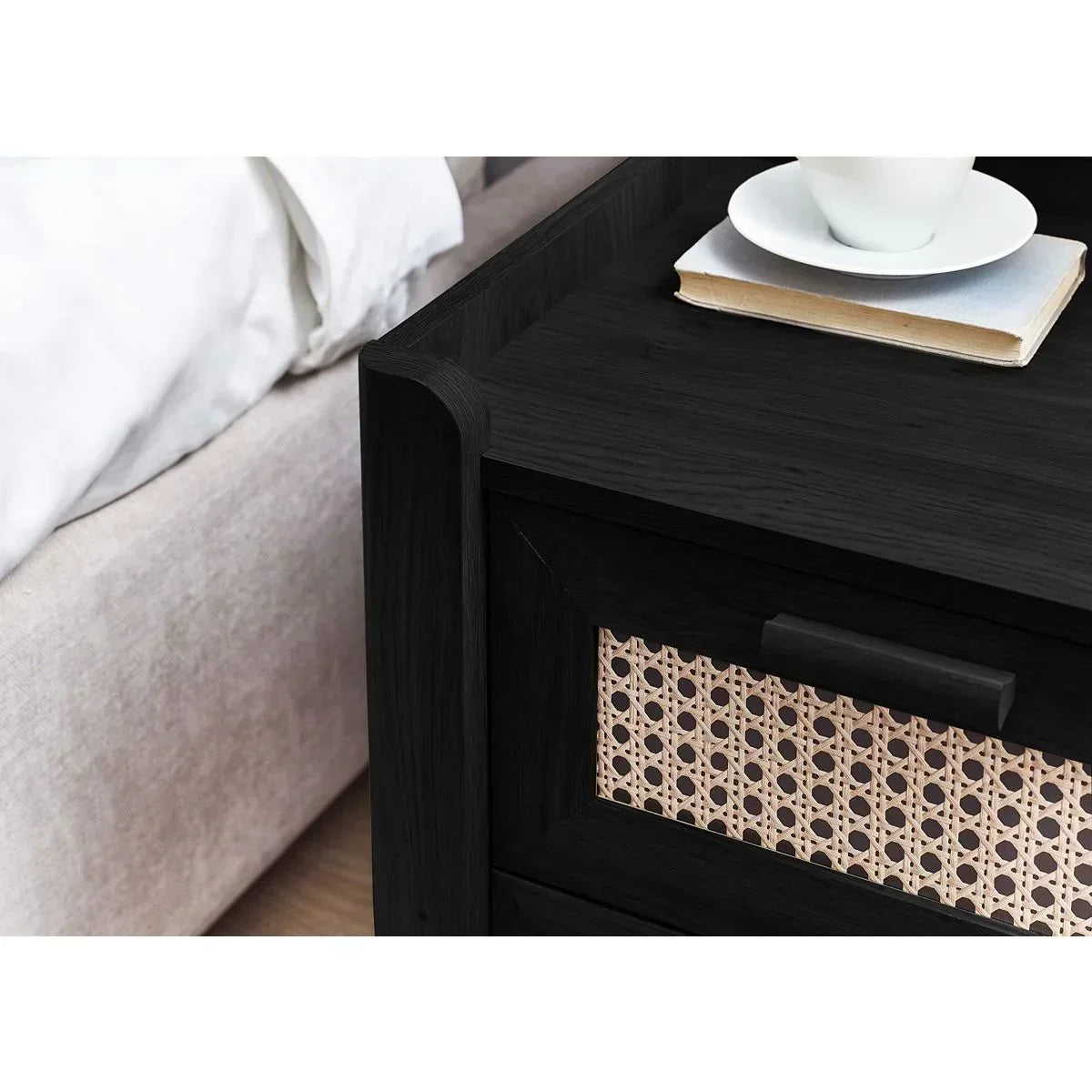 Black wooden bedside table with cane drawer front beside bed, cup and book on top