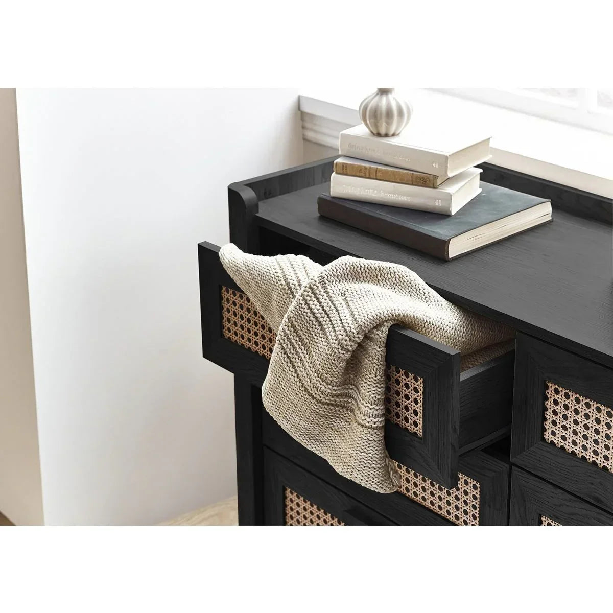 Black wood and rattan chest of drawers with open drawer, beige throw, and stacked books