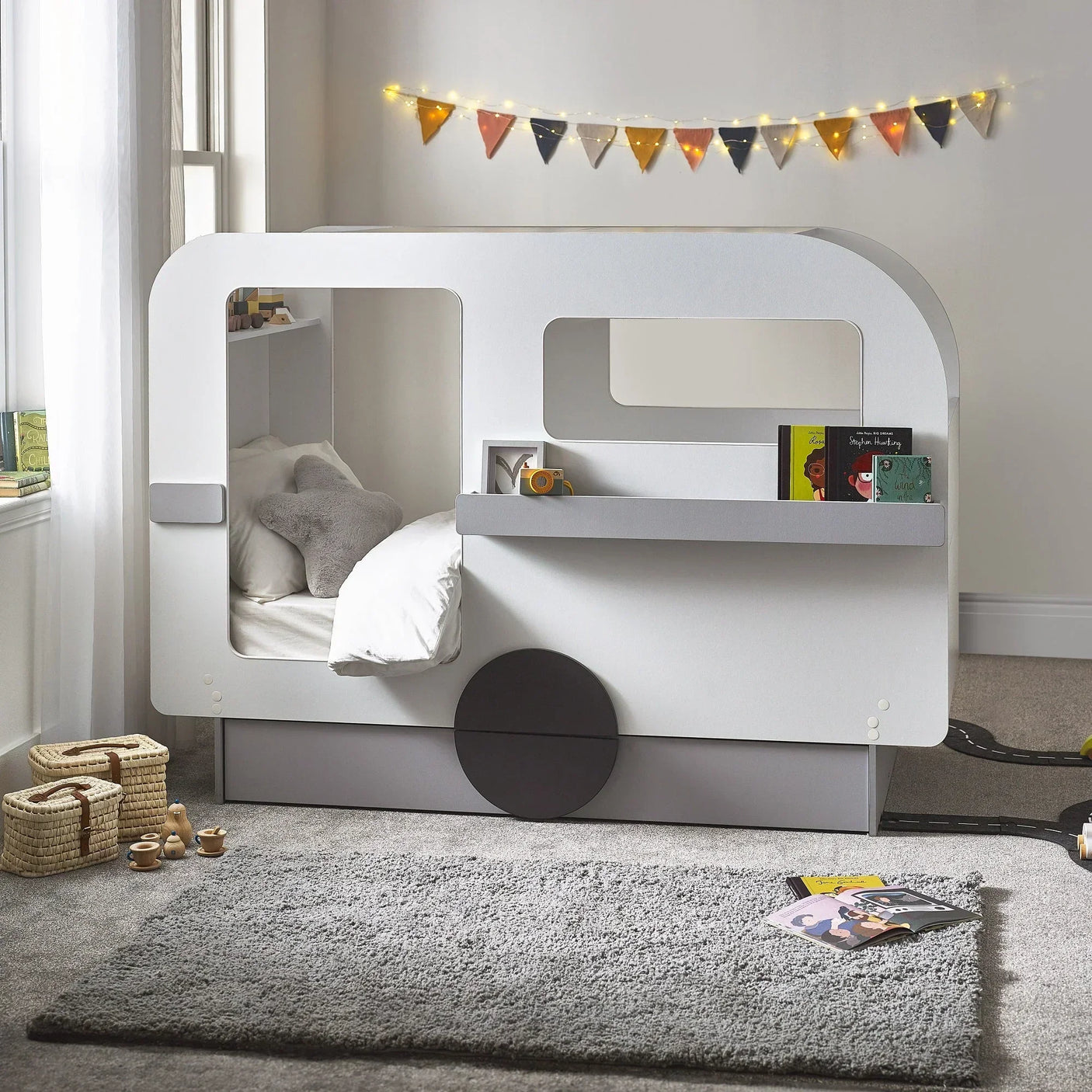 Caravan shaped kids bed with window cut-outs, built-in shelf, and books, in bright bedroom