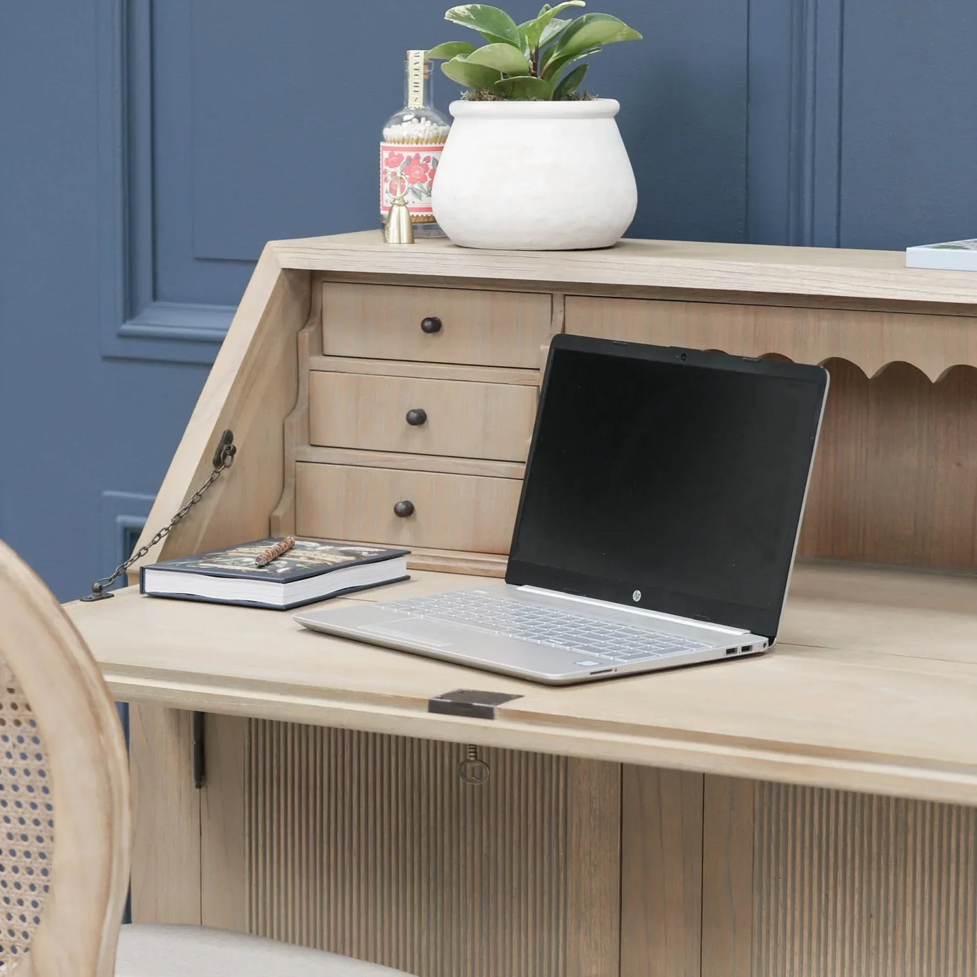 Wooden writing desk with laptop, book, potted plant and drawers against blue wall
