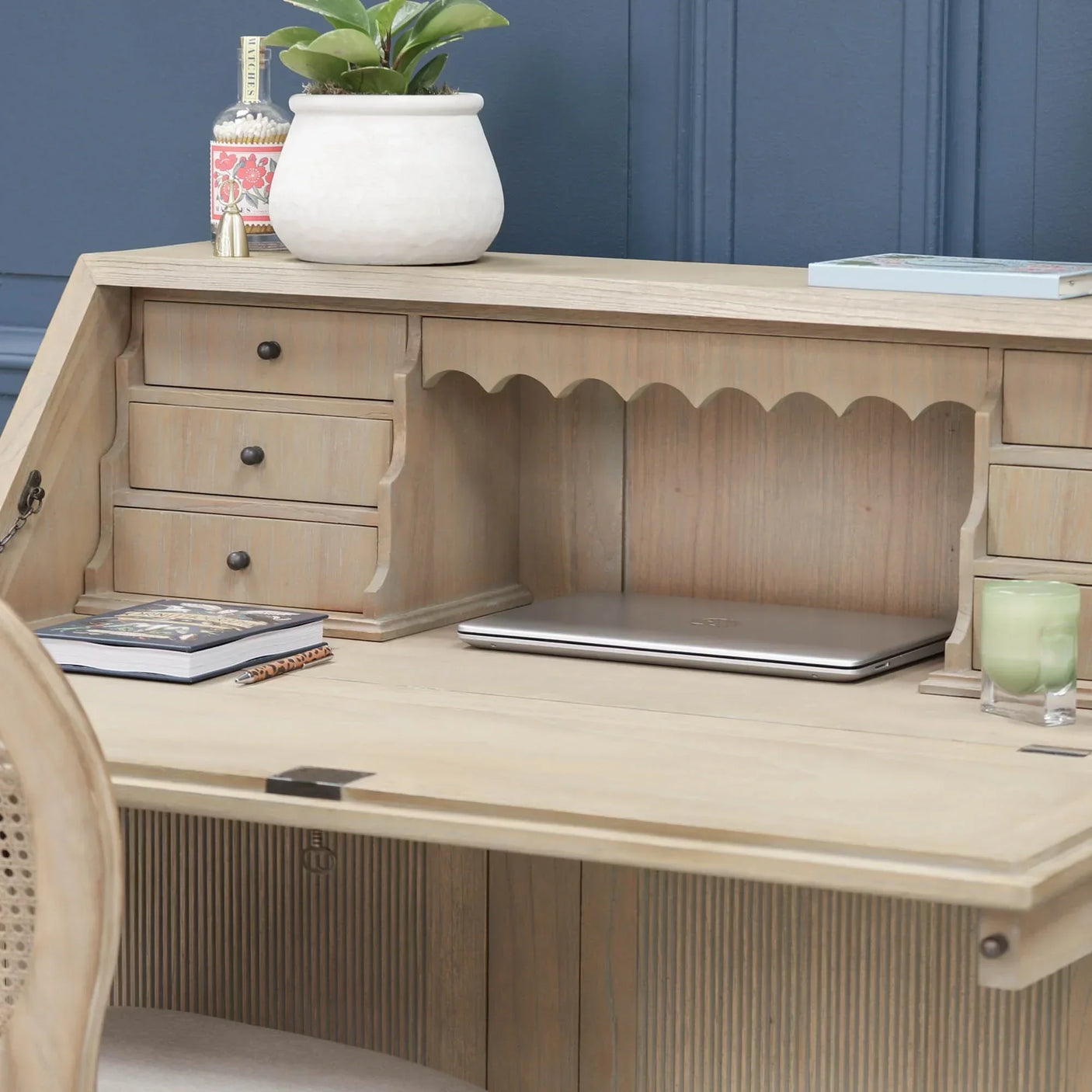 Wood writing desk with drawers, fold-down front, and decorative plant against blue wall
