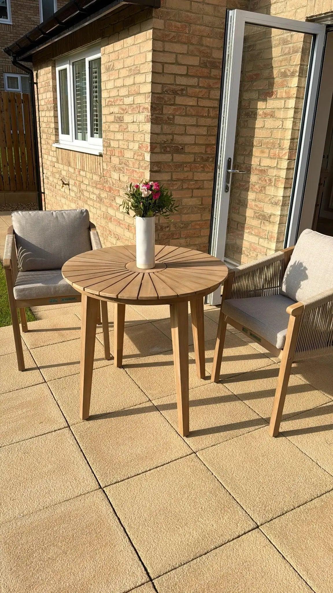 Wooden outdoor dining set with round table, two cushioned chairs, vase of flowers on patio