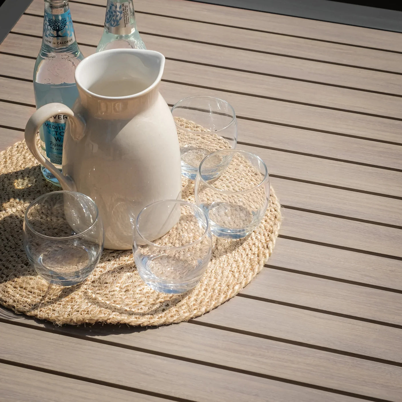 Light wood outdoor dining table with jug, glass tumblers, and water bottles on woven mat