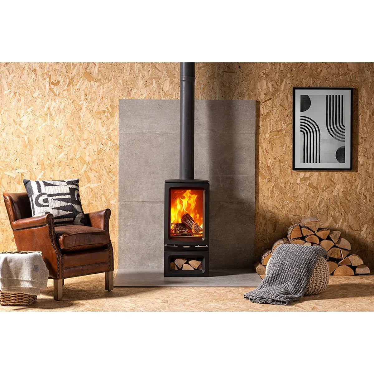 Wood burning stove with fire, stacked logs, leather armchair, and modern wall art