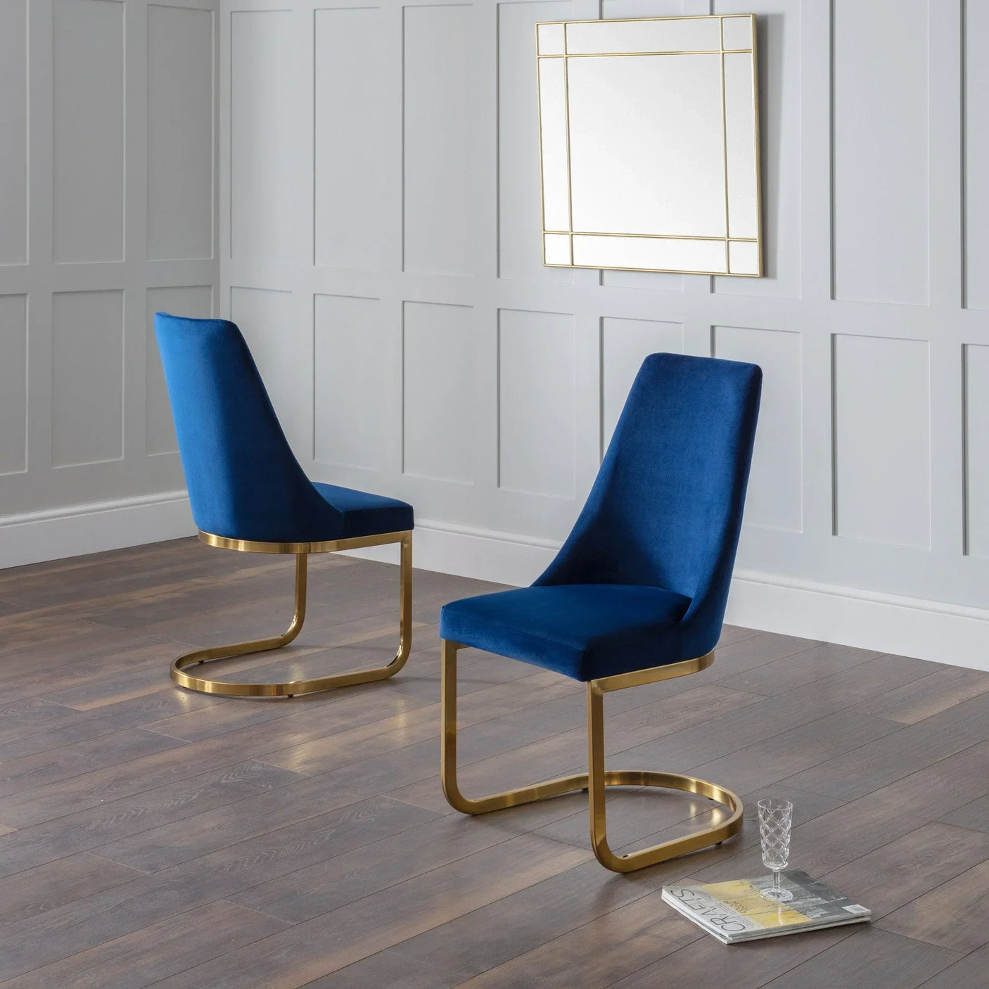 Blue velvet dining chairs with gold cantilever frames on wood floor, glass and magazine nearby