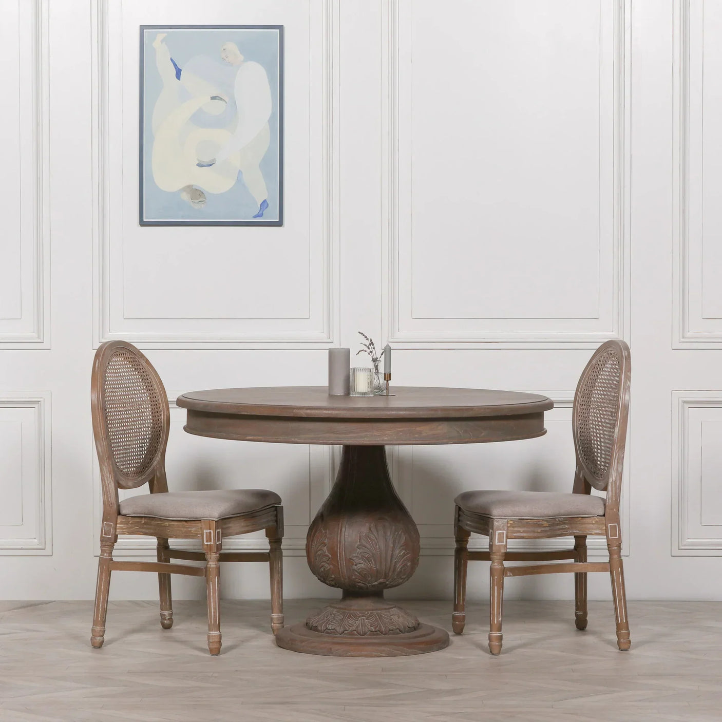 Round wooden dining table with pedestal base and two cane back chairs in neutral room