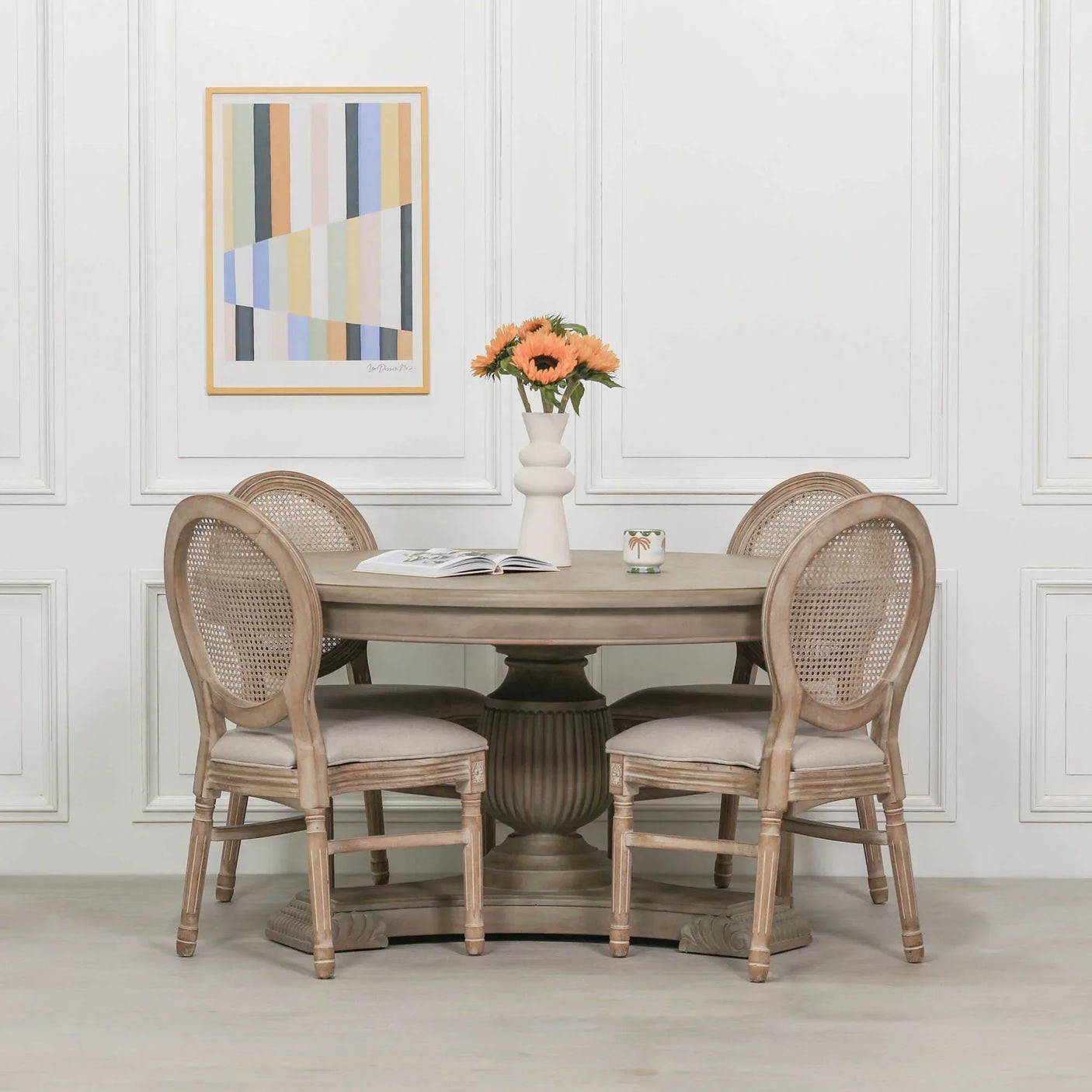 Round wooden dining table with four cane back chairs in neutral room, vase of flowers on top
