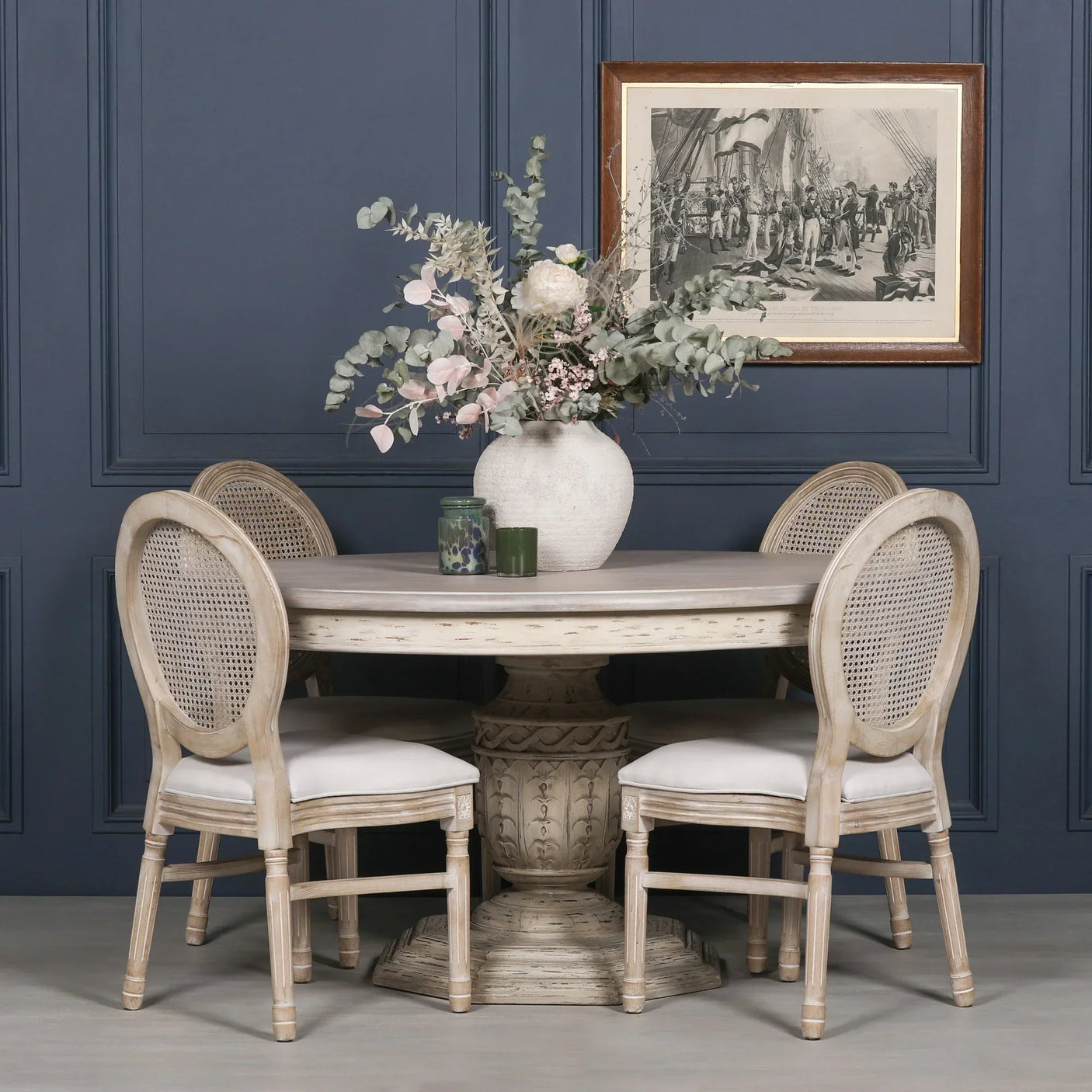 Round wooden dining table with carved base and four rattan back chairs in neutral finish