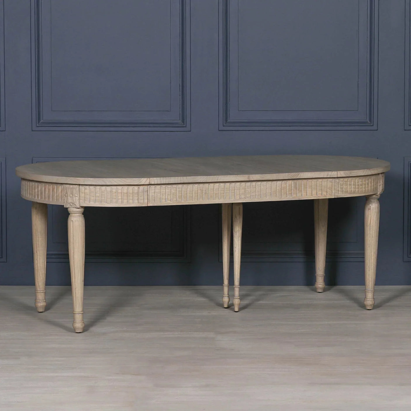 Oval wooden dining table with carved apron and six tapered legs against dark blue wall
