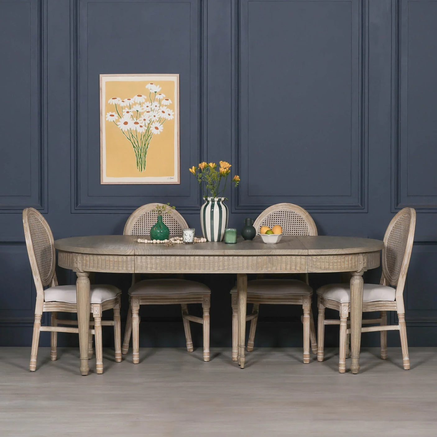 Oval wooden dining table with four rattan back chairs in front of navy panelled wall
