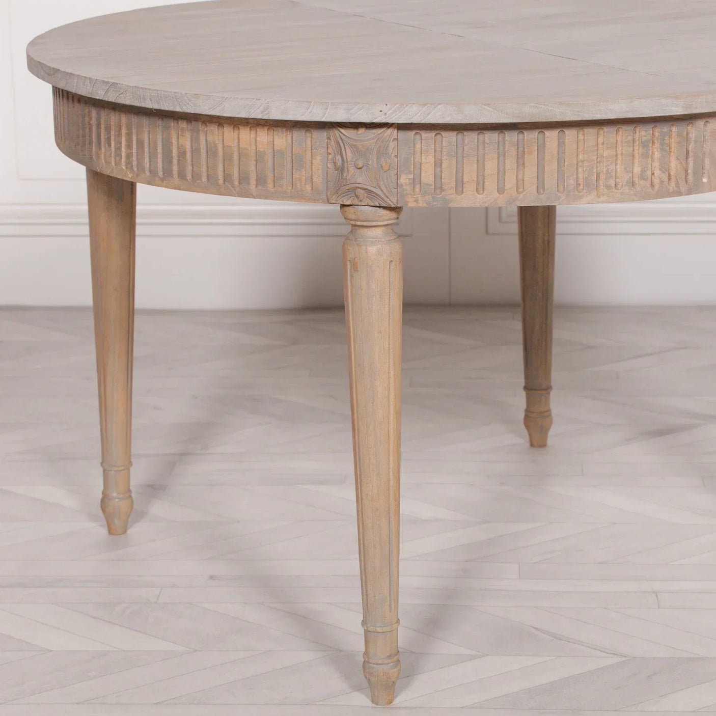 Round wooden dining table with carved detailing and tapered legs on light herringbone floor
