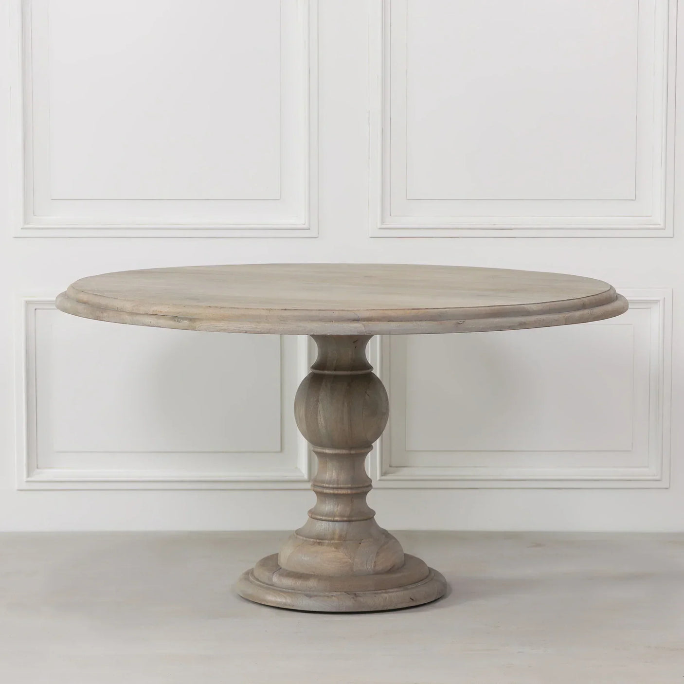 Round wooden dining table with pedestal base in light oak finish against panelled wall