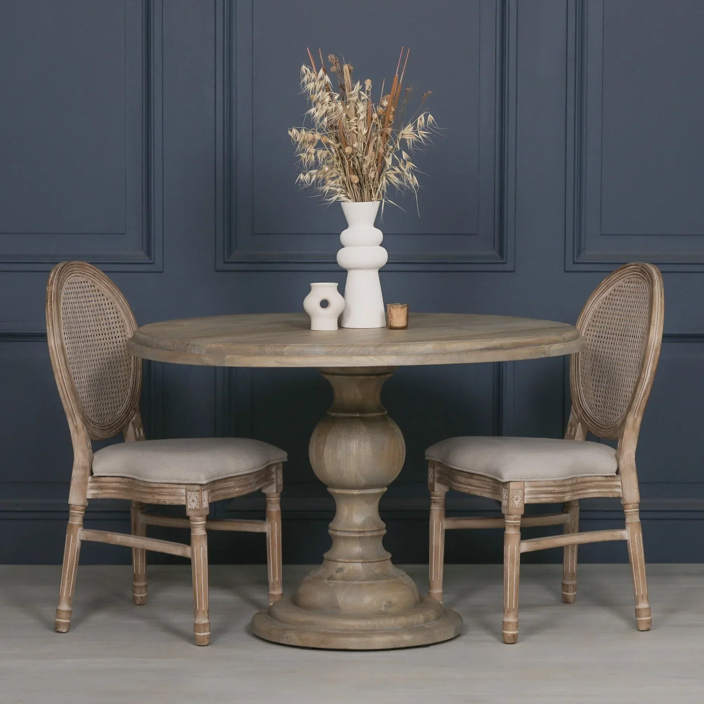 Round wooden dining table with pedestal base and two upholstered chairs, dried flower vase