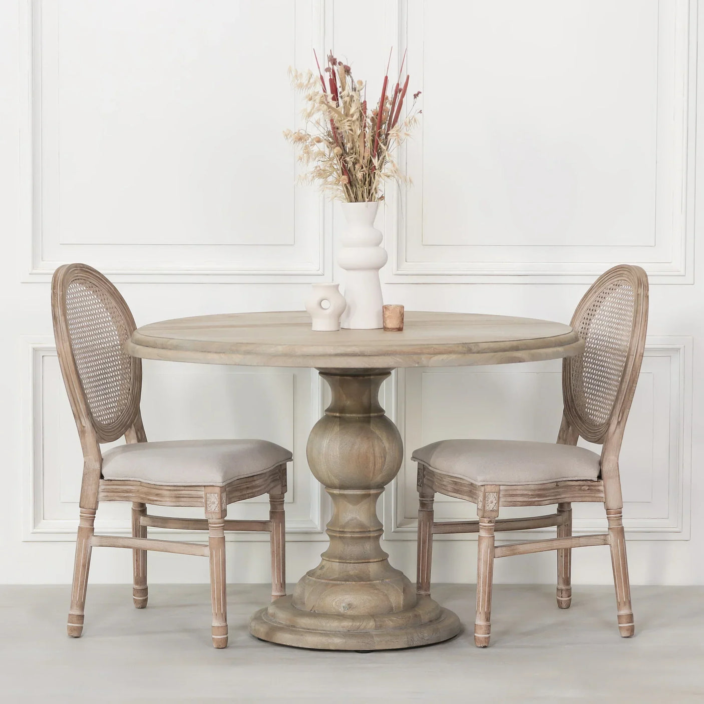 Round wooden dining table with pedestal base and two upholstered chairs in neutral tones