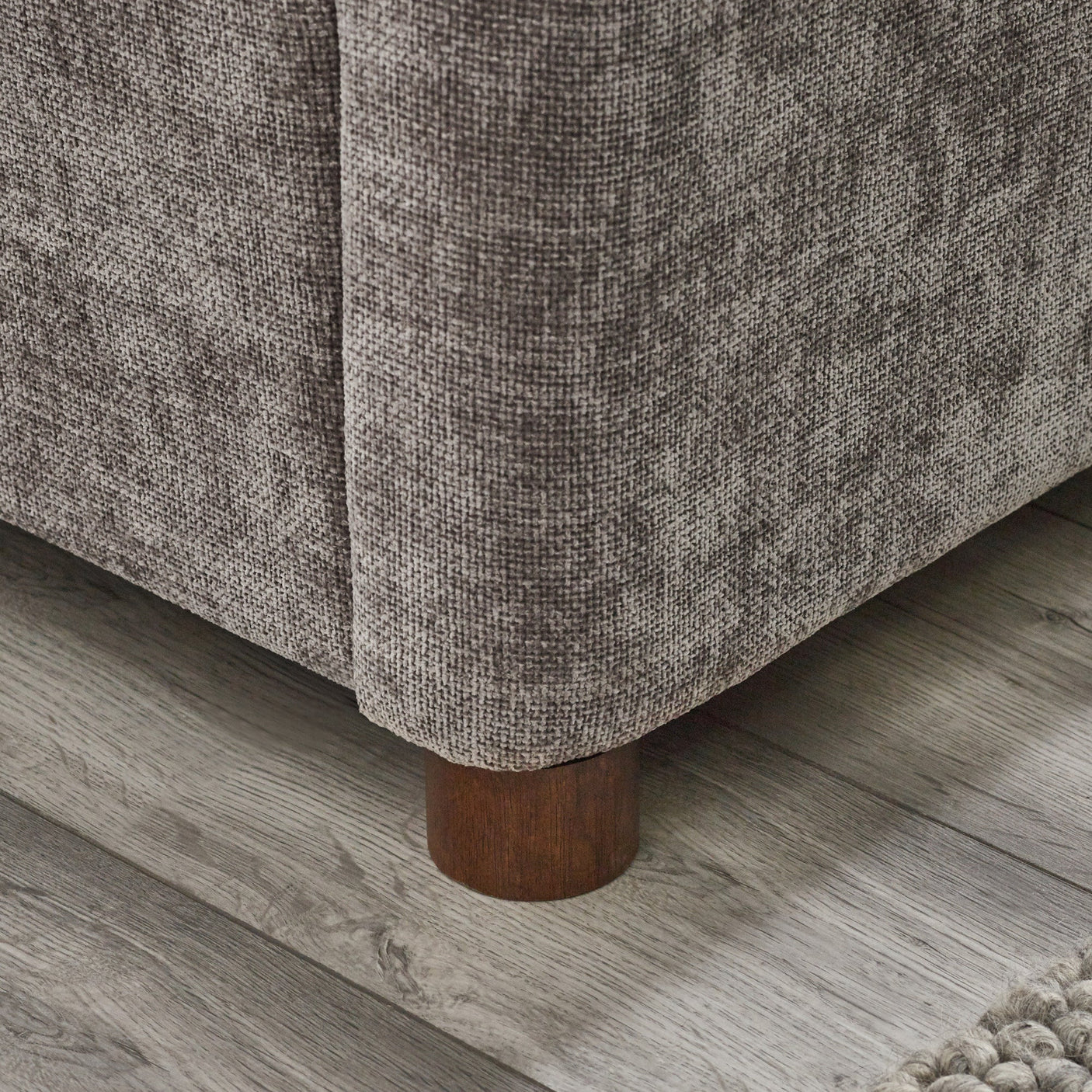 Close-up corner view of rounded wooden bed leg against wood floor and grey upholstery
