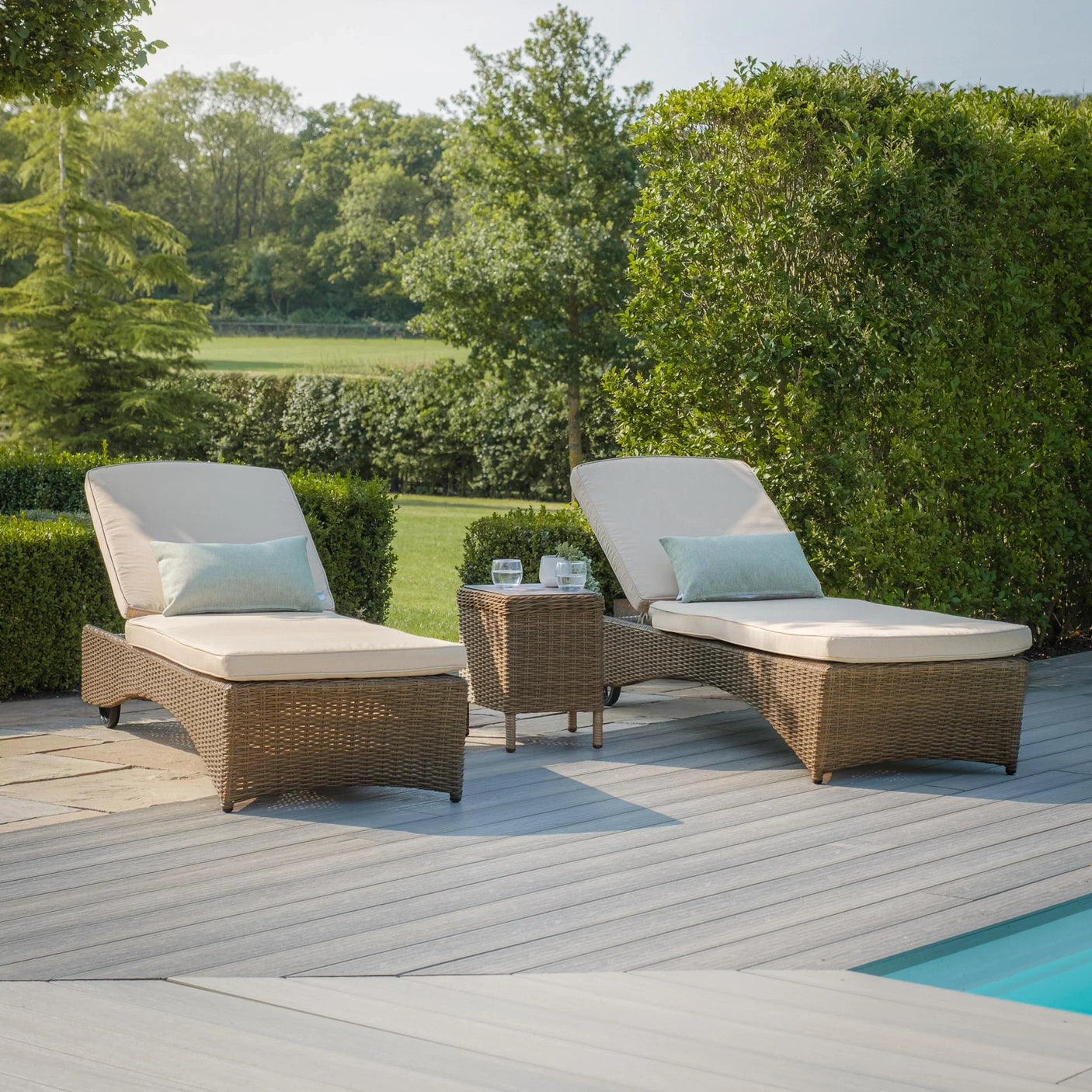 Rattan sun loungers with cream cushions and side table by pool on patio