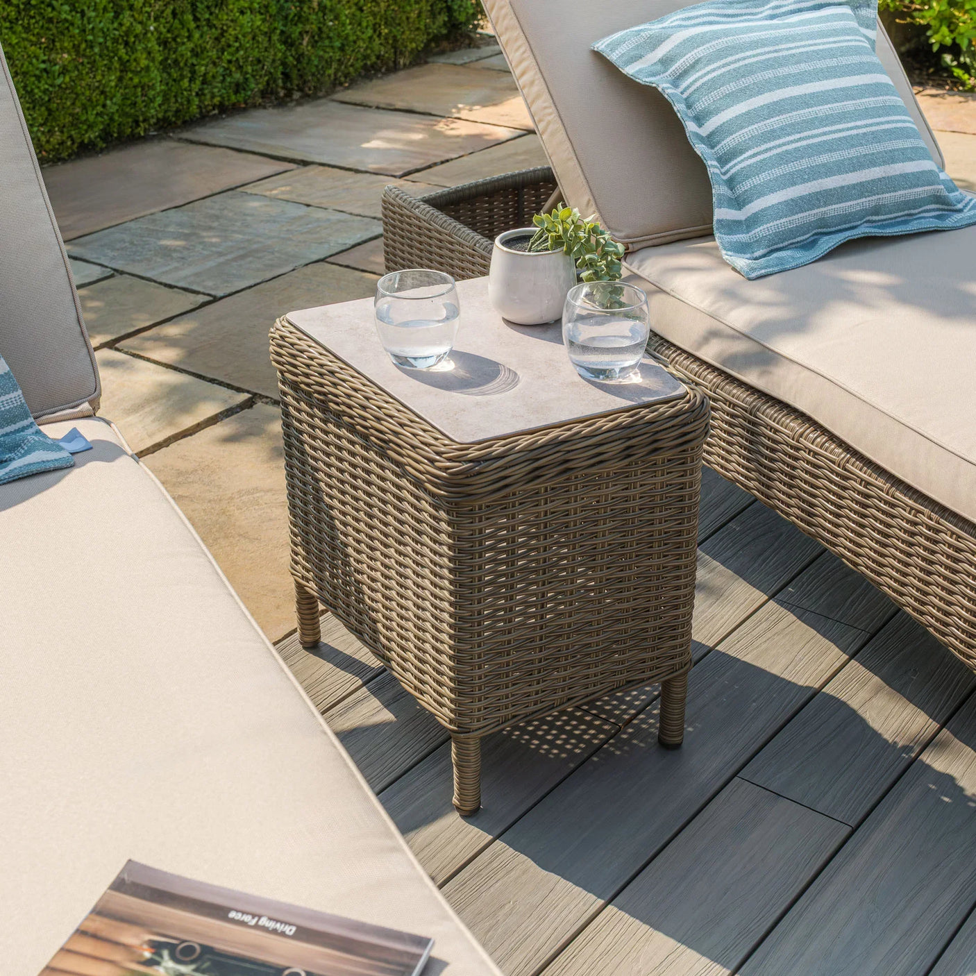 Rattan outdoor side table with glass top between two beige garden loungers on patio deck