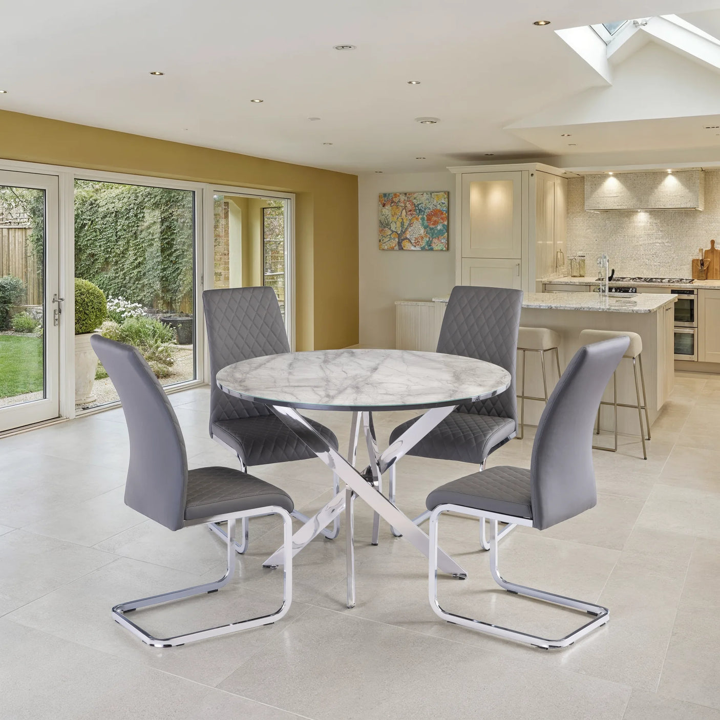 A modern round dining table with a white and gray marble-patterned top and chrome cross base, surrounded by four gray upholstered chairs with chrome cantilever legs, set in a bright contemporary kitchen and dining room.