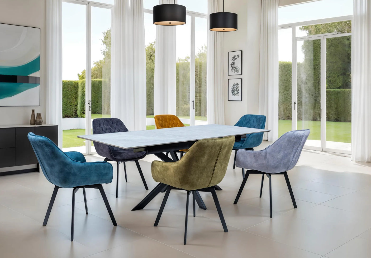 A modern dining room scene with a rectangular white dining table surrounded by six upholstered chairs in blue, gray, yellow, and purple velvet, set in a bright room with large windows and outdoor views.
