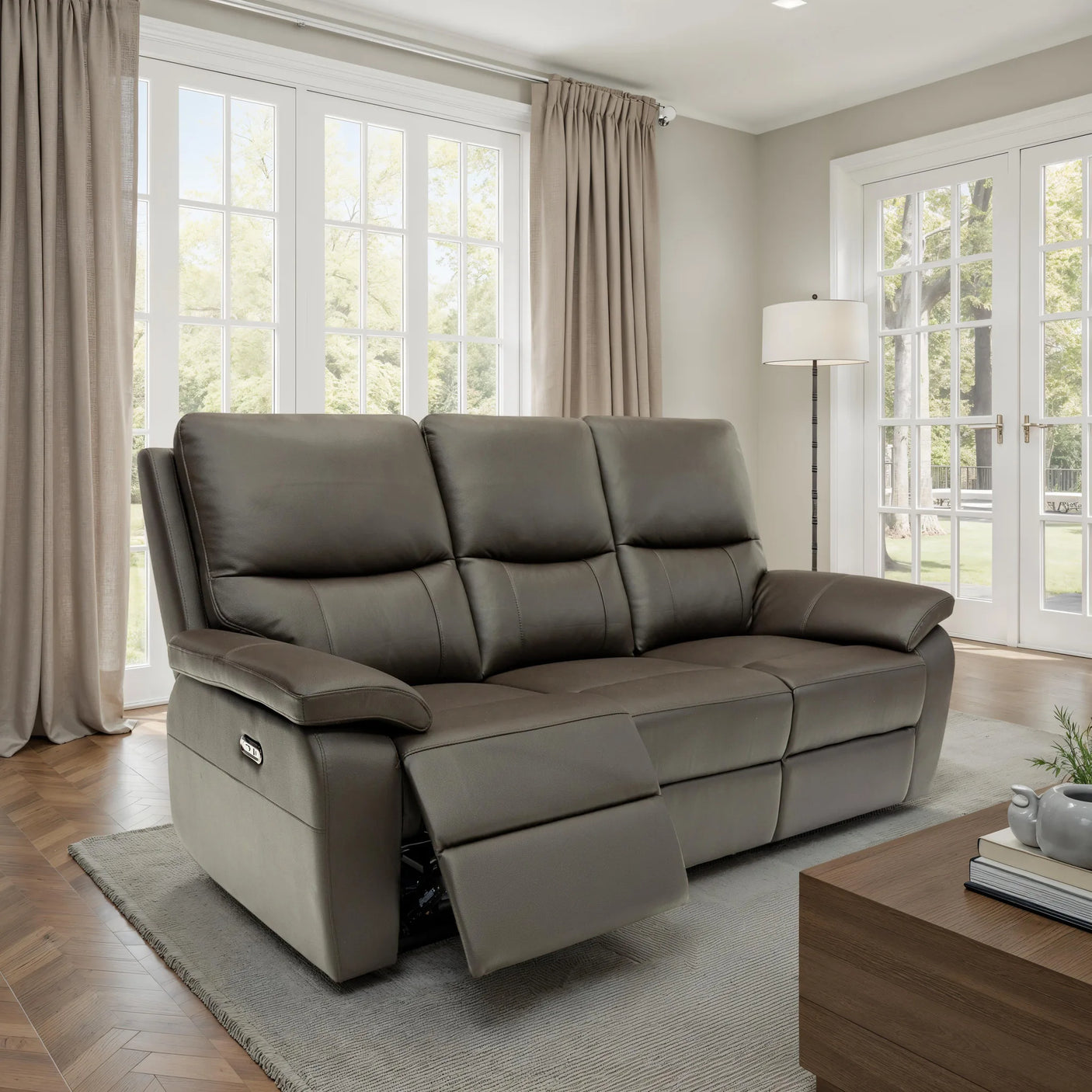 A three-seat reclining leather sofa in taupe brown, shown with the front left cushion extended, set in a bright modern living room with large windows, beige curtains, and a wooden coffee table.