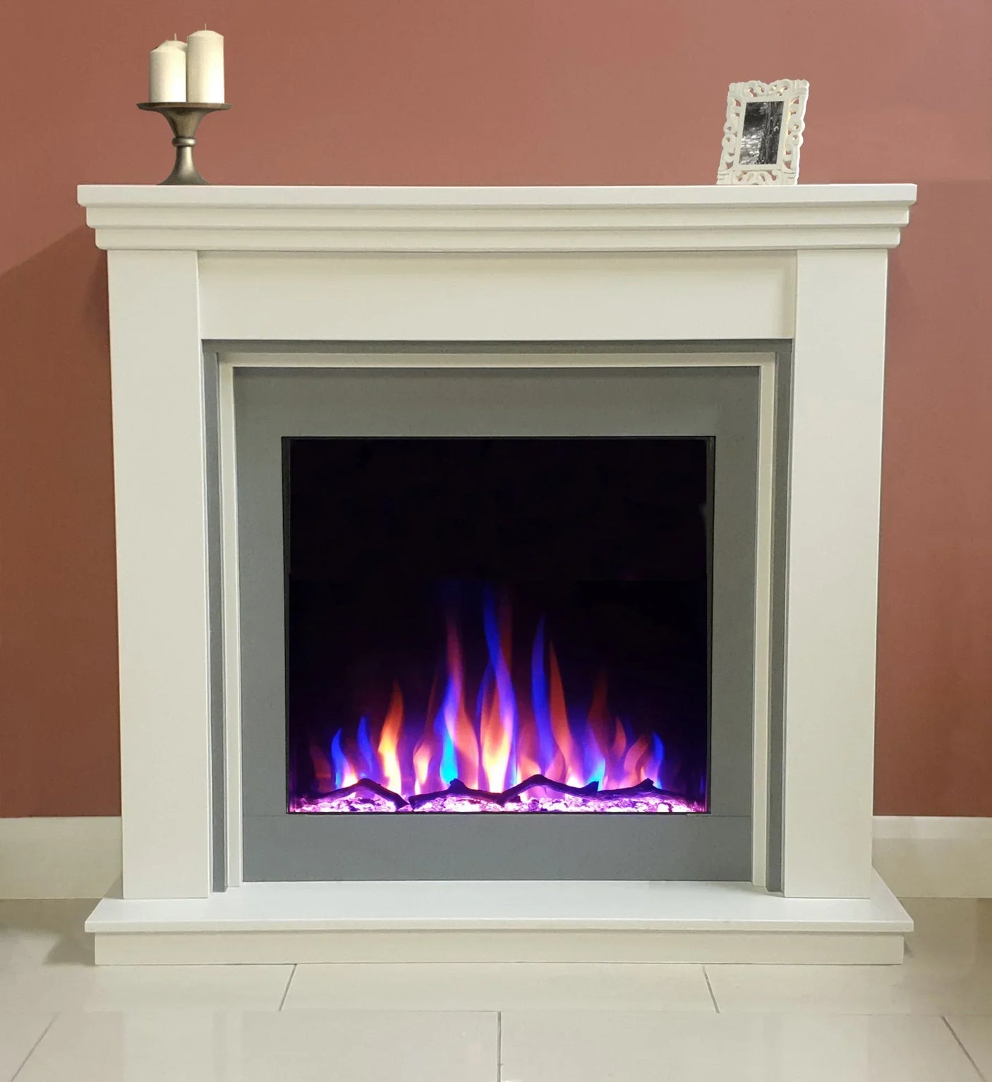 White electric fireplace with multicolored LED flames against pink wall, candles on mantel